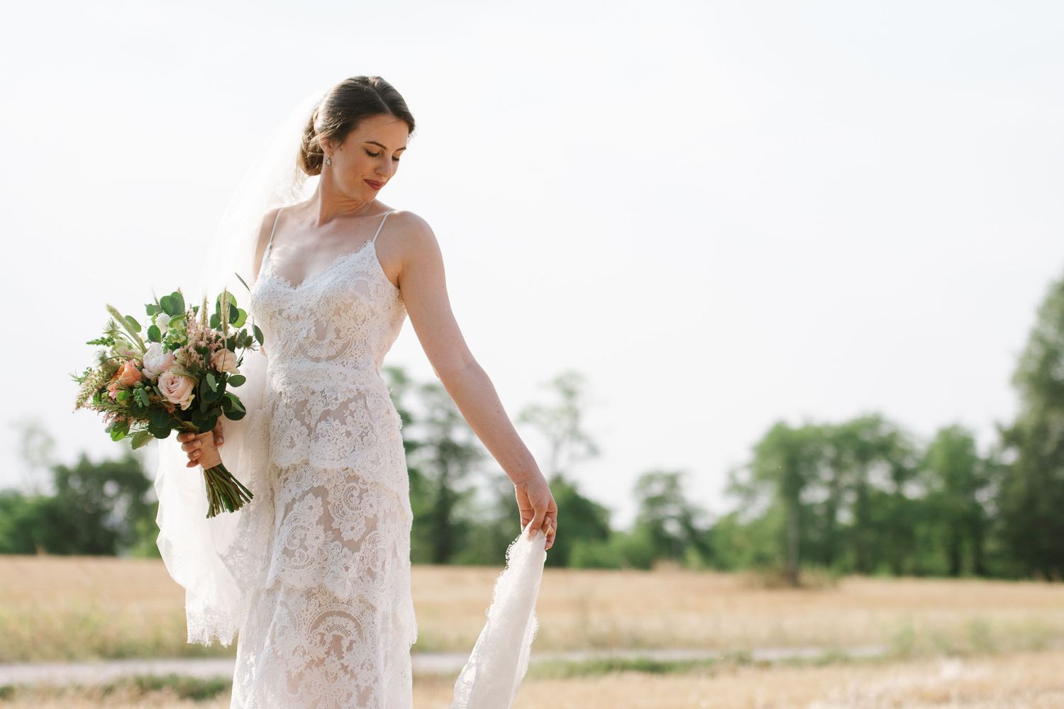 A bride in a detailed lace wedding gown holds her bouquet of peach and white flowers in a sunlit field.