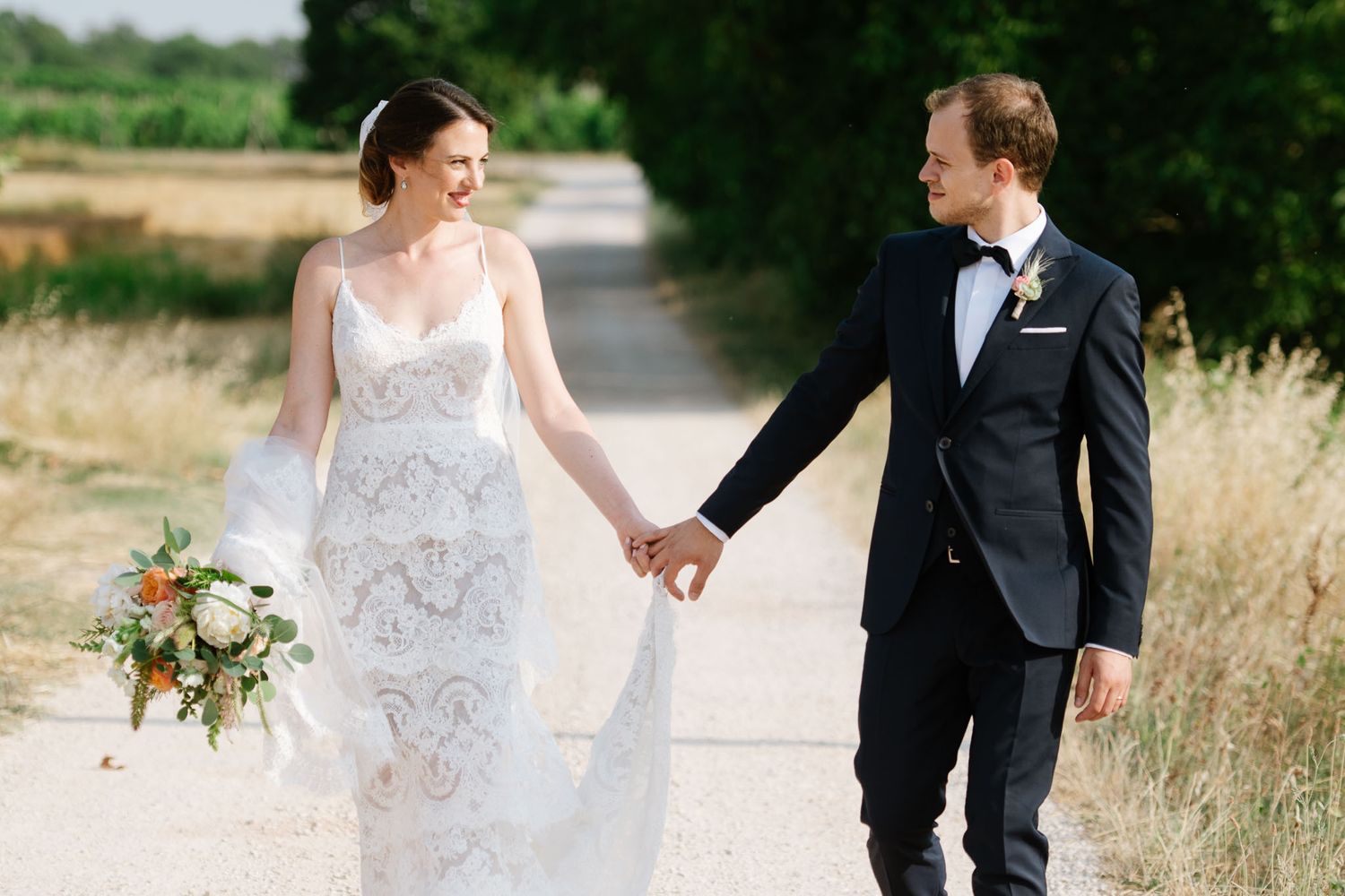 A bride and groom hold hands while walking down a countryside path surrounded by tall grass.