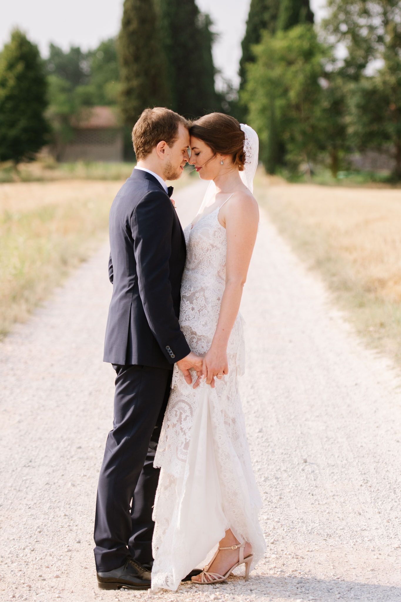 A tender moment between newlyweds on a white gravel path with cypress trees in background.