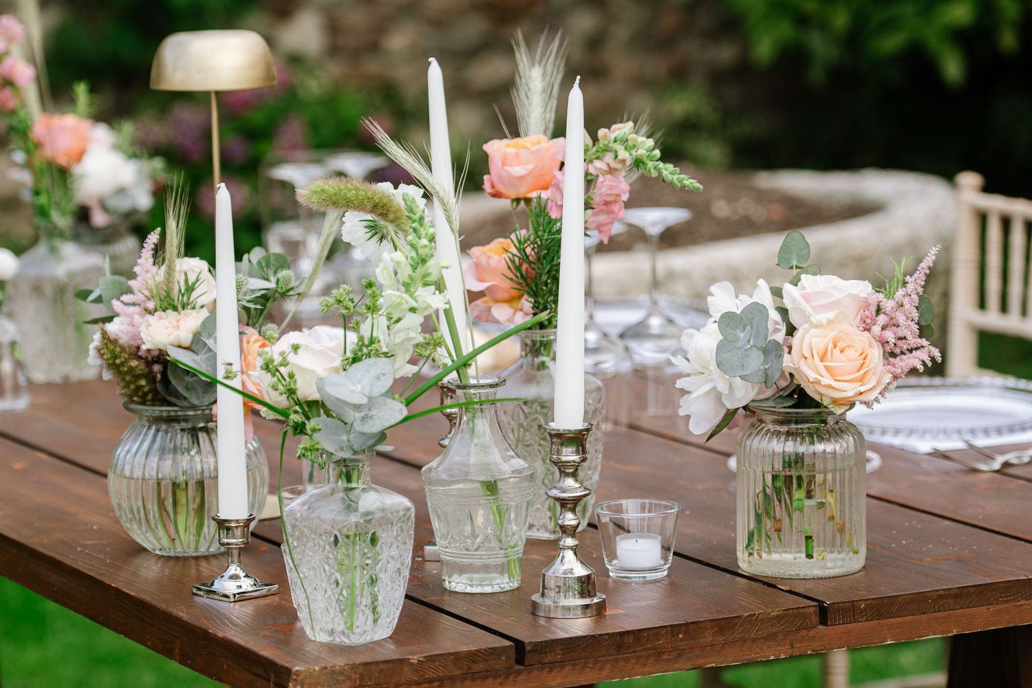 Rustic wedding table decor with glass vases, pink flowers and white candles on wooden surface.