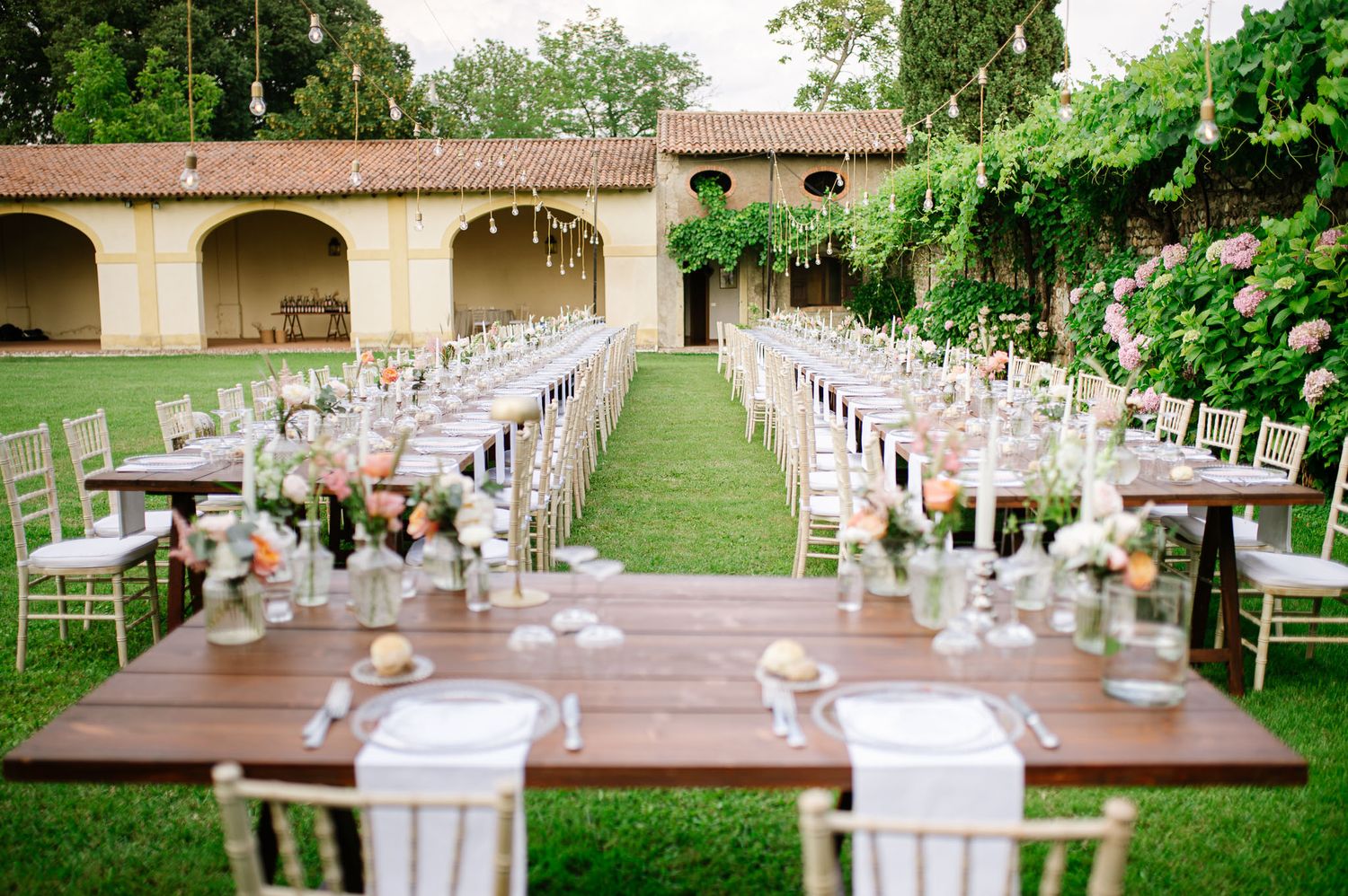An outdoor wedding reception setup with long wooden tables and white chairs in a Mediterranean courtyard.