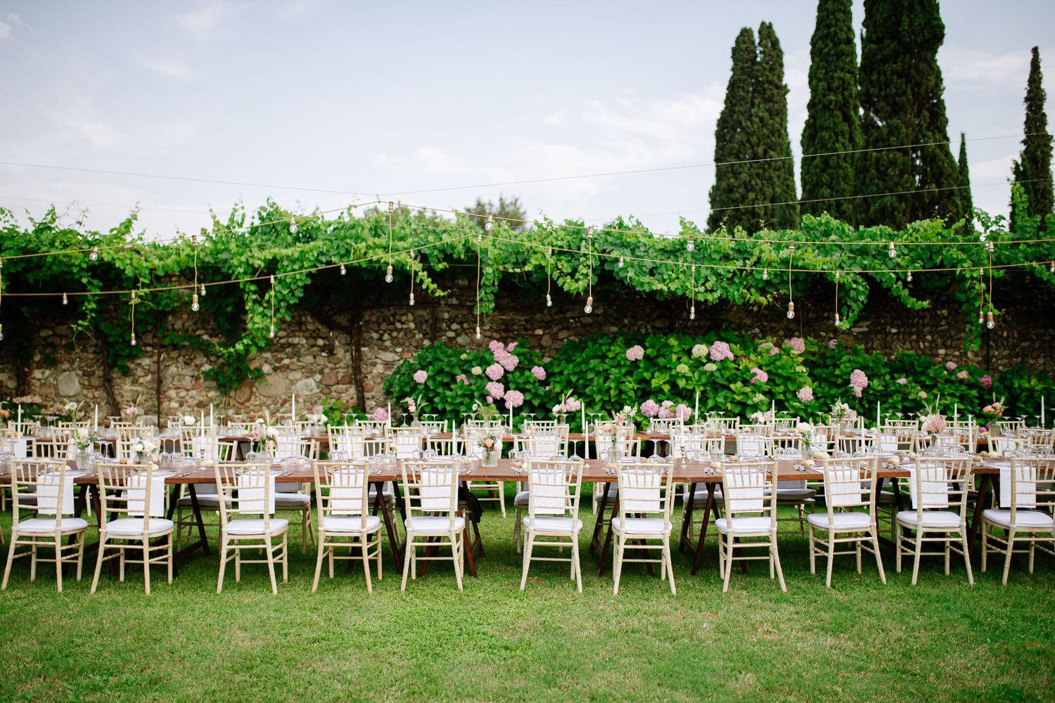Elegant outdoor wedding reception table setup with white chairs along a stone wall covered in lush greenery and cypress trees.