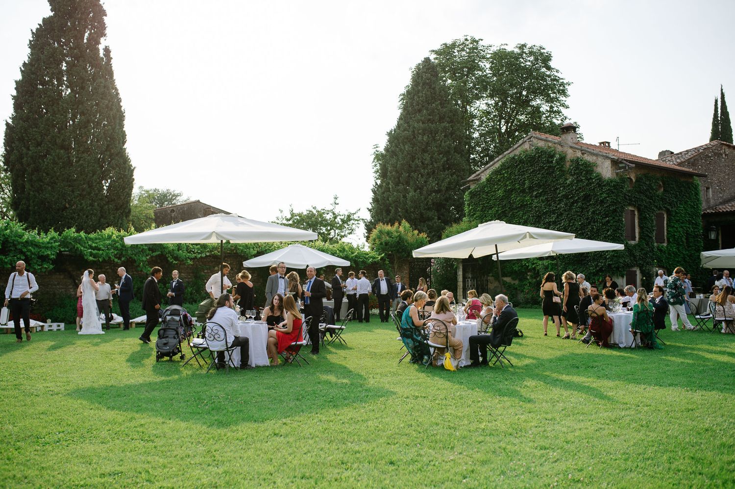 Outdoor garden reception with white umbrellas and guests seated at tables on a manicured lawn.