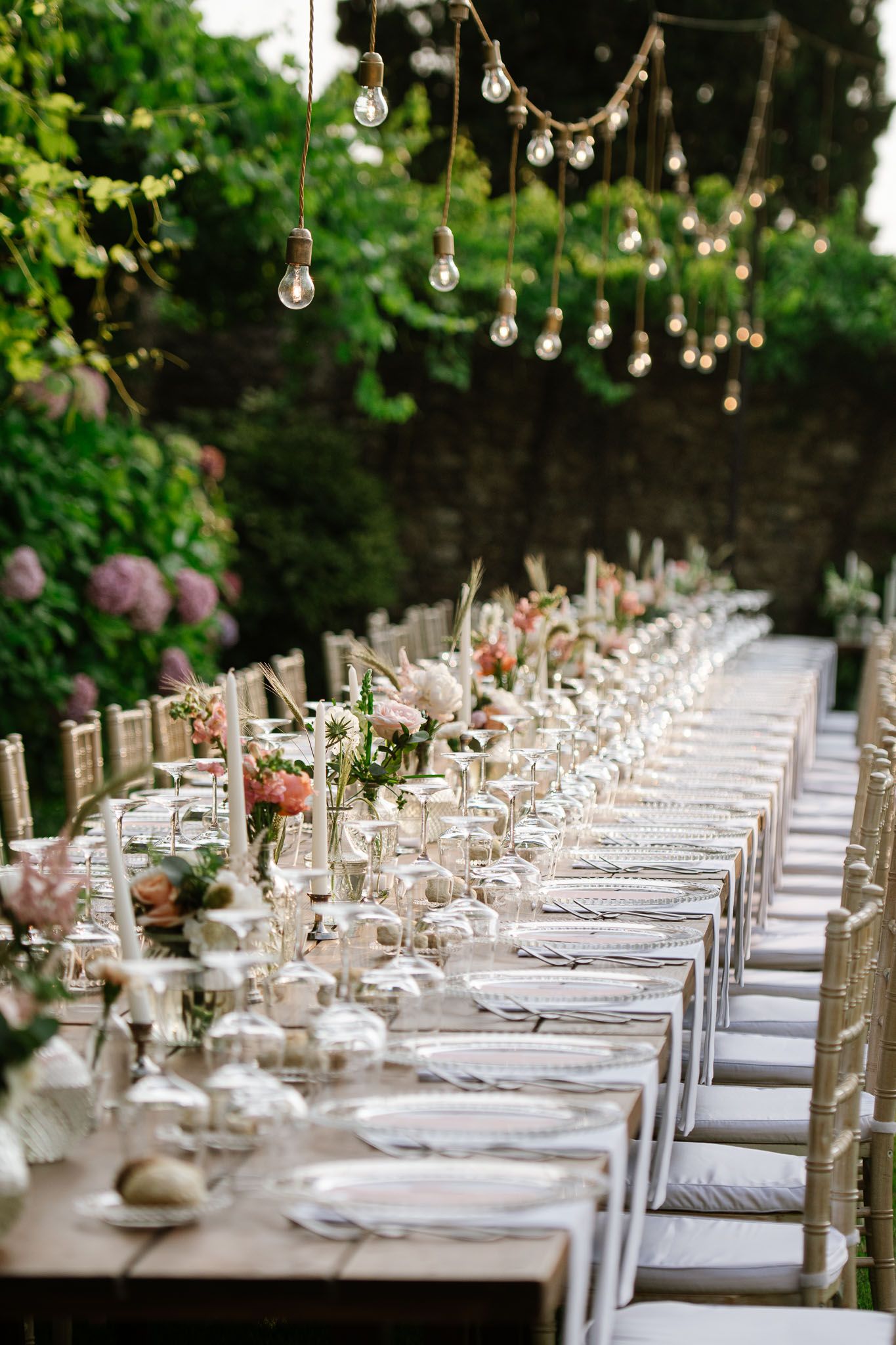 Long wedding reception table with hanging lights, white place settings and pink floral centerpieces.
