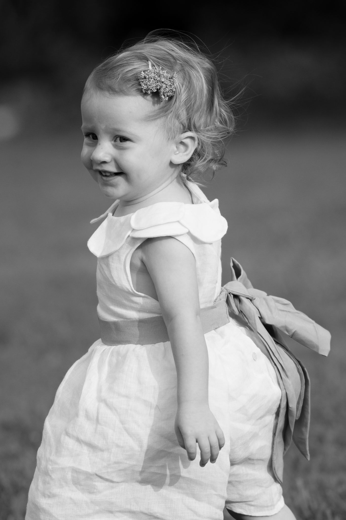 A sweet toddler in a white dress with a delicate hair accessory smiles.