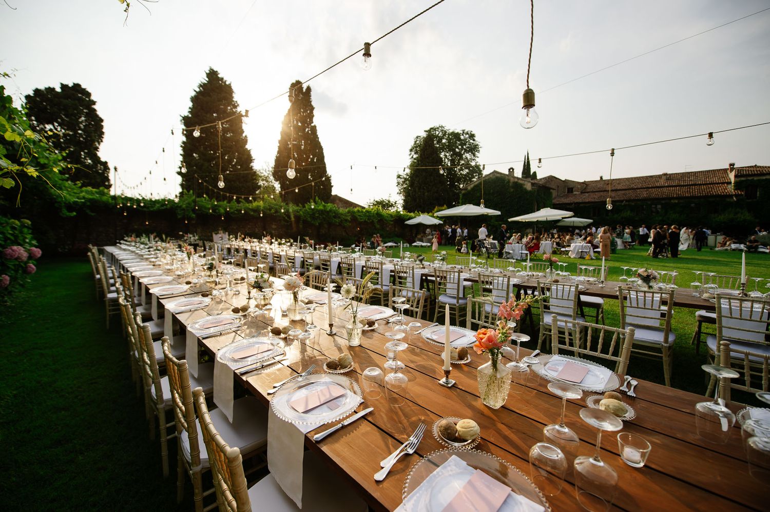 Rustic outdoor wedding reception tables set under string lights with white chairs and table settings.