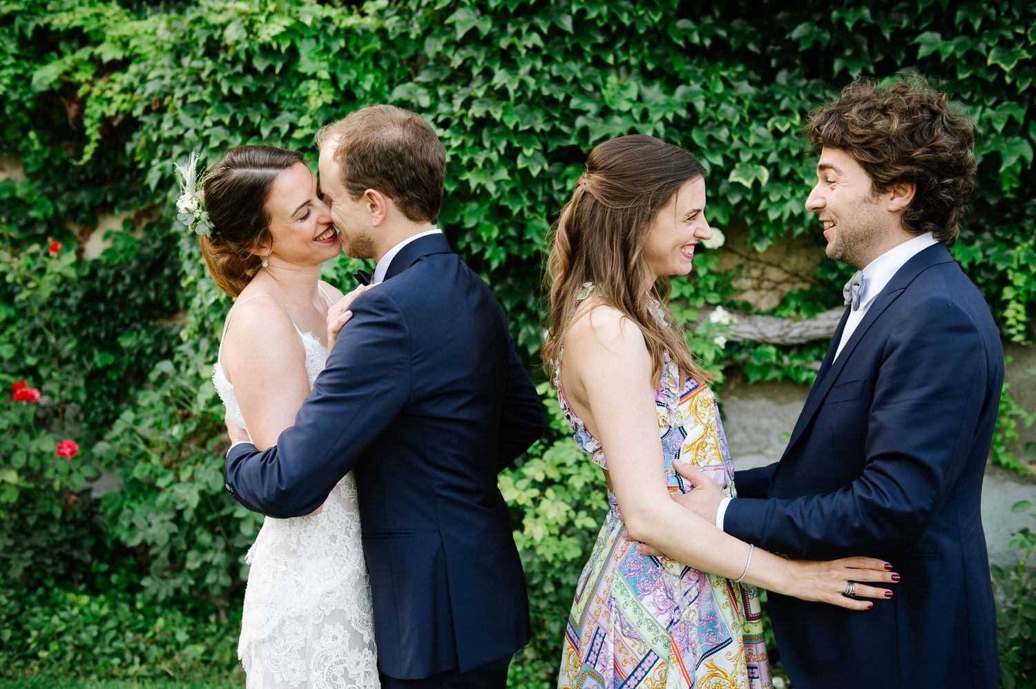 Two couples share intimate moments in a lush garden setting during wedding celebrations.