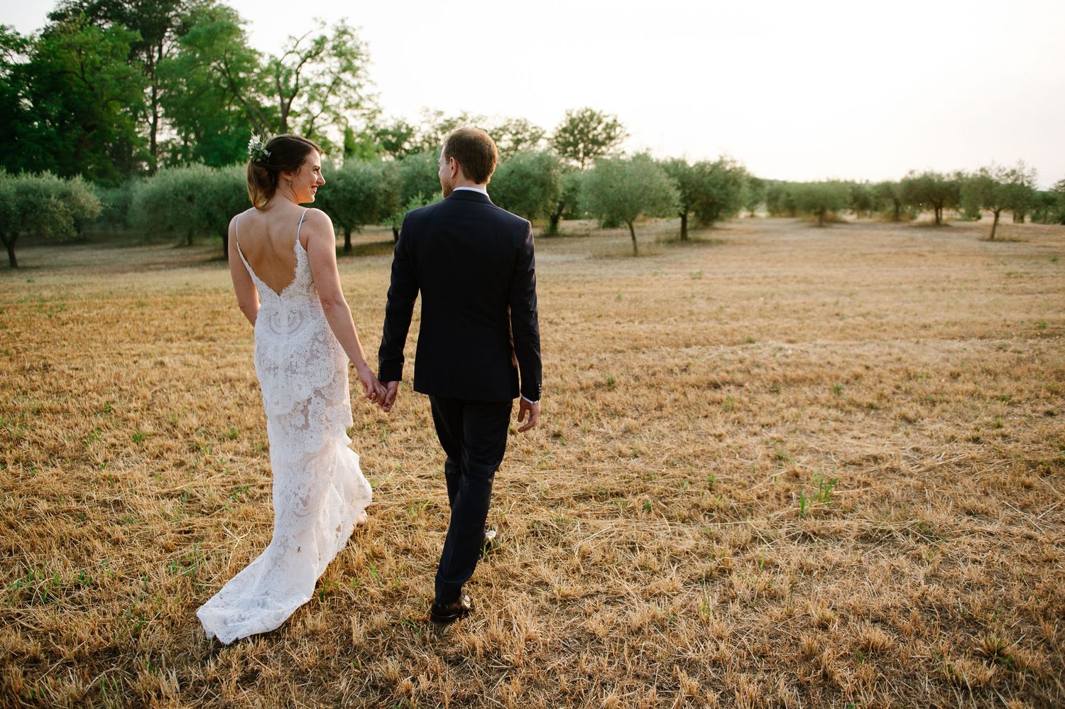 Newlyweds walking hand in hand through dry grass field at sunset.