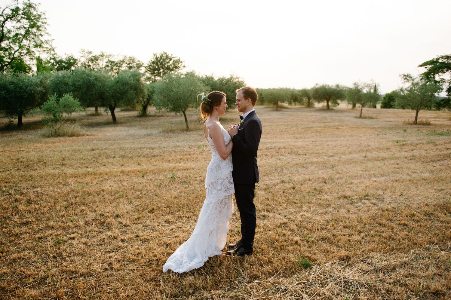 Bride and groom embrace in golden sunlit field surrounded by scattered trees.