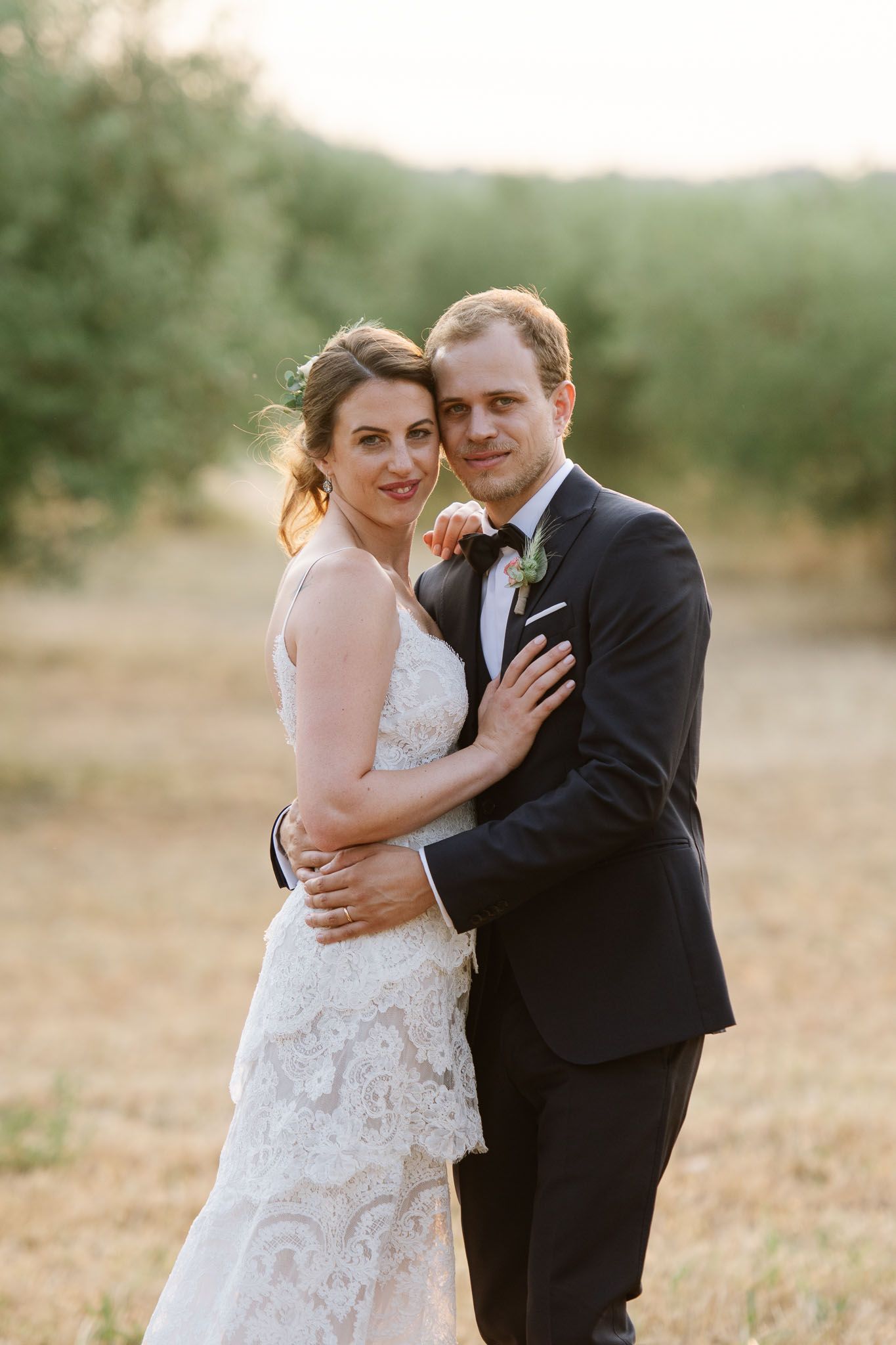 Bride in lace gown and groom in black suit pose in golden hour lighting.