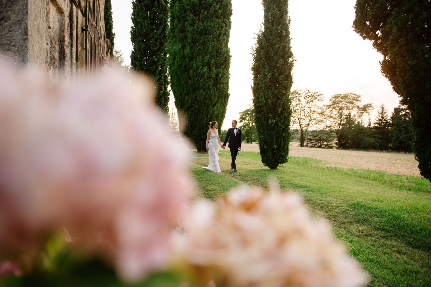 A couple walks along a cypress-lined path at sunset with pink flowers in the foreground.