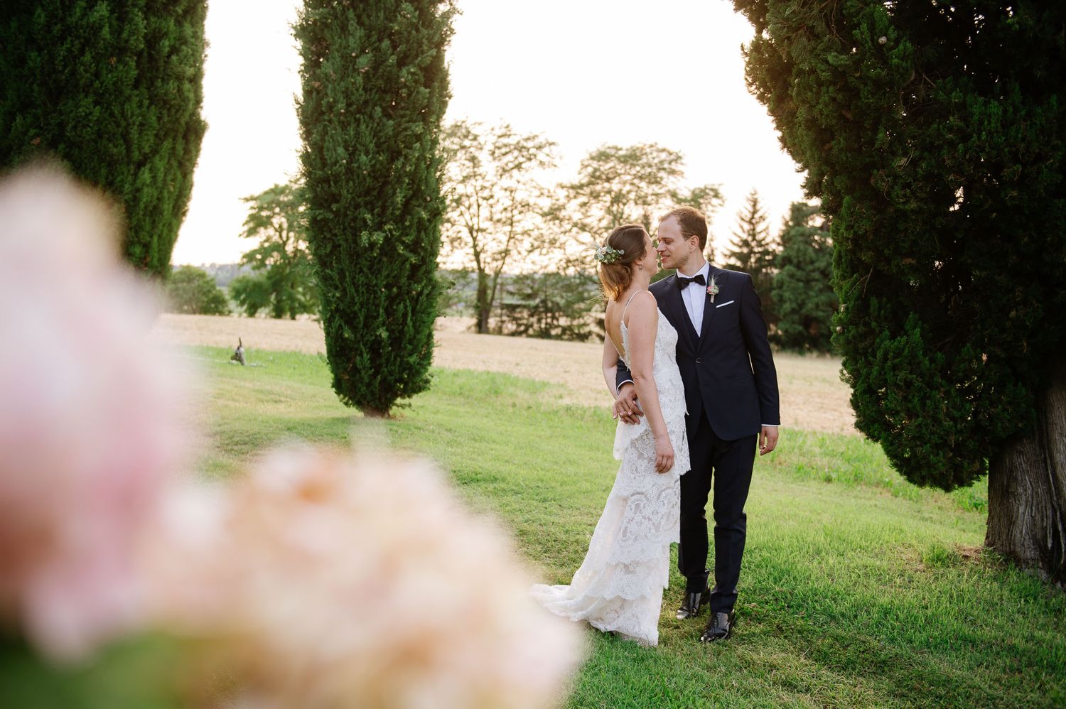 Elegant wedding couple poses between towering cypress trees in a garden setting.