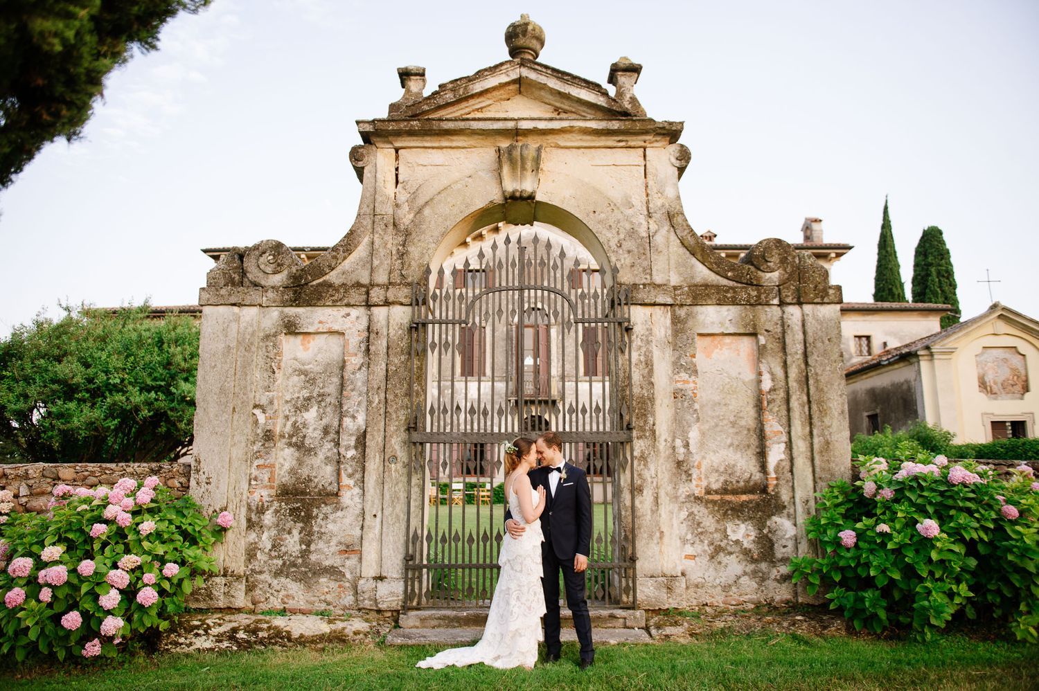 Wedding couple poses in front of a grand historic stone archway with roses.