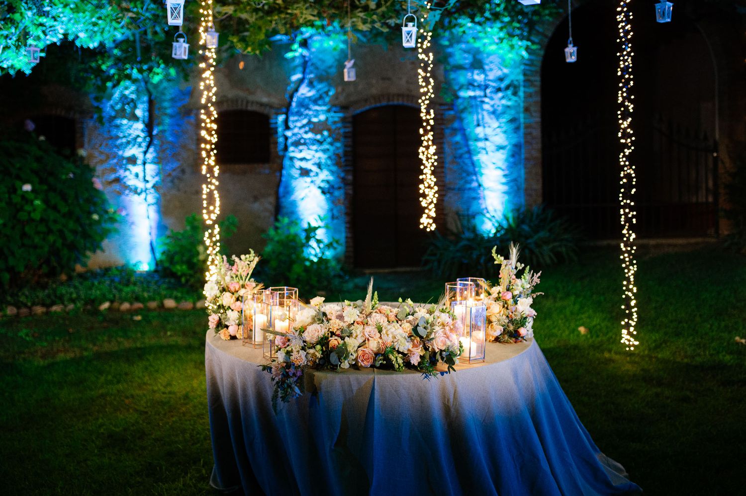 Romantic reception table with hanging lights and blue uplighting in garden setting.