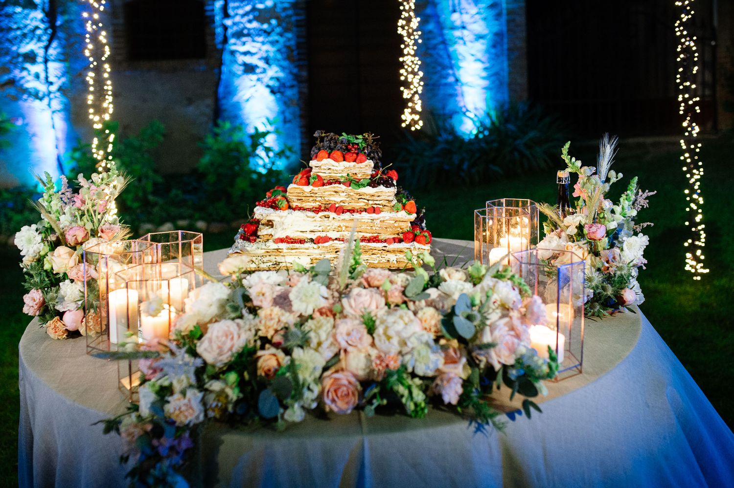 Elegant wedding cake display with cascading flowers and romantic blue uplighting in an outdoor evening setting.