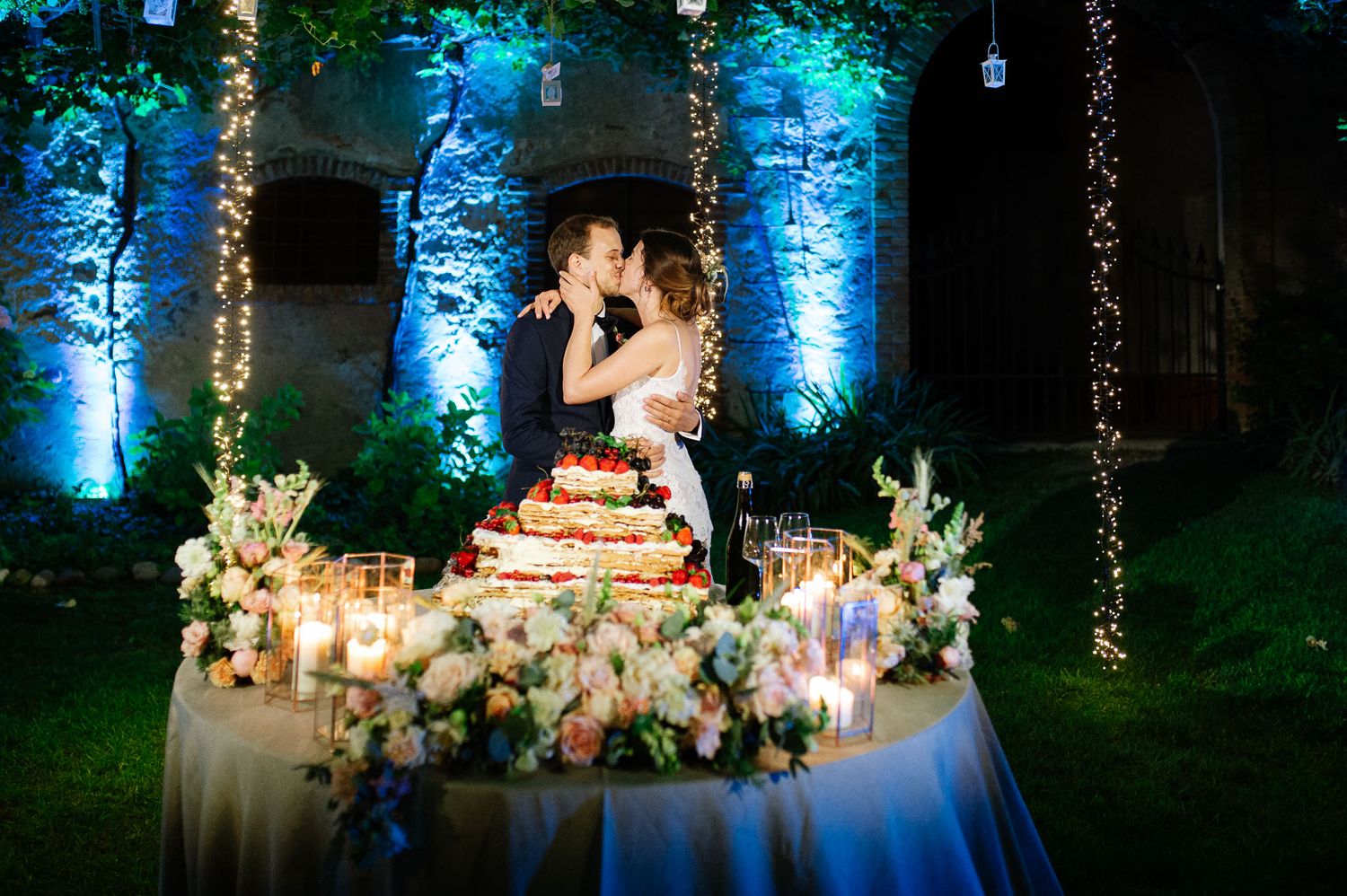 Wedding cake table decorated with lush floral arrangements and string lights creating a magical nighttime atmosphere.