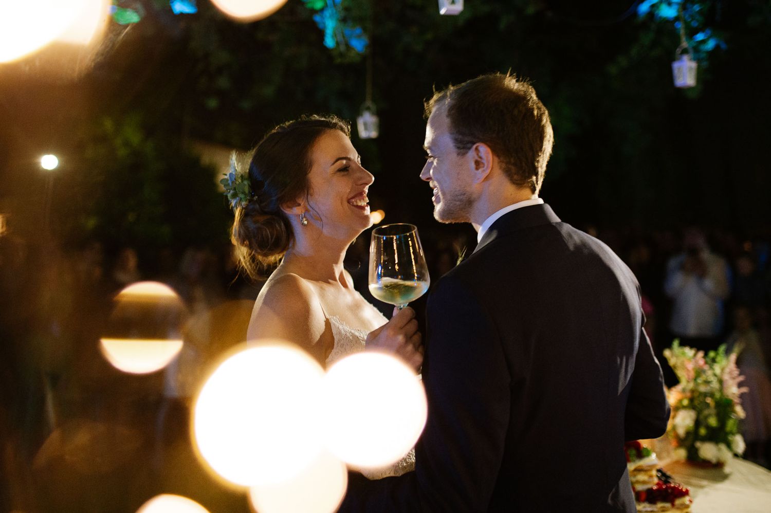Romantic nighttime wedding reception scene with wine glasses and atmospheric bokeh lighting effects.