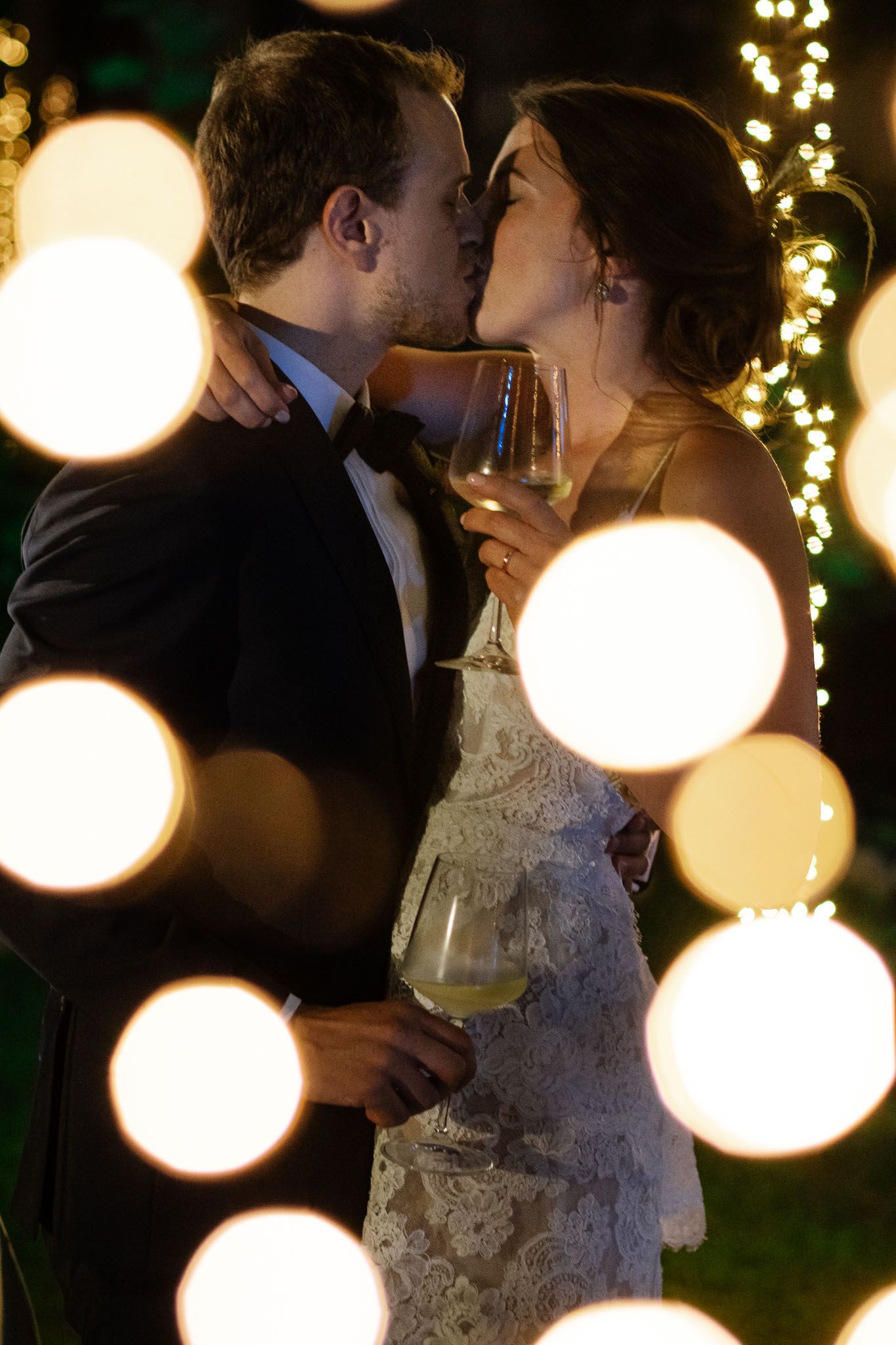 Intimate wedding moment captured with dreamy bokeh lights and wine glasses in an evening garden setting.
