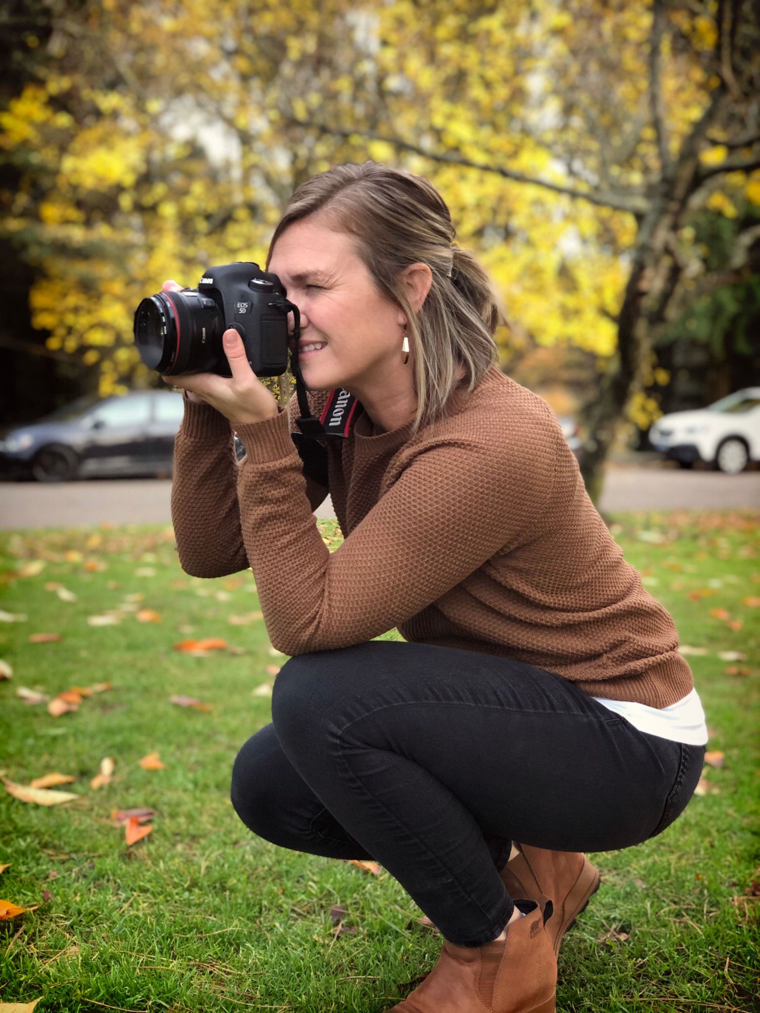 Woman photographer crouching outdoors with DSLR camera in autumn park with colorful fall foliage.
