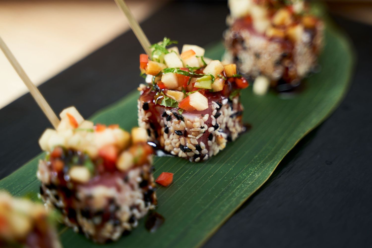 Sushi rolls coated in black sesame seeds served on a banana leaf with colorful toppings.