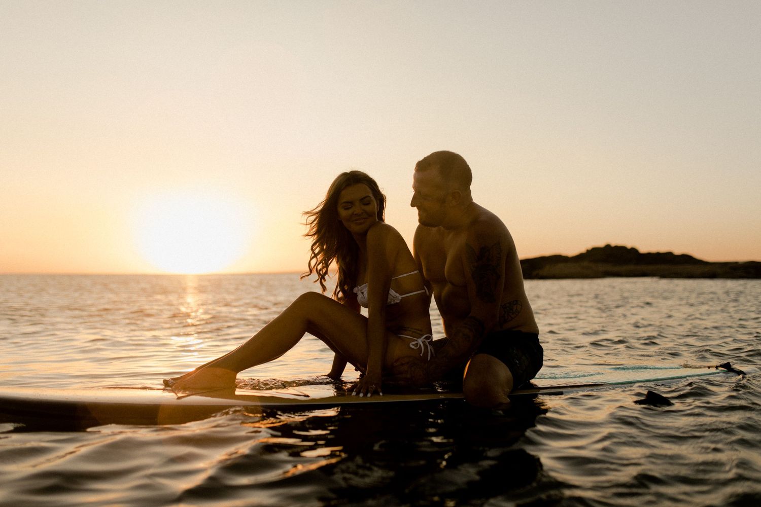 Couple sitting together on a surfboard during golden hour sunset at sea.