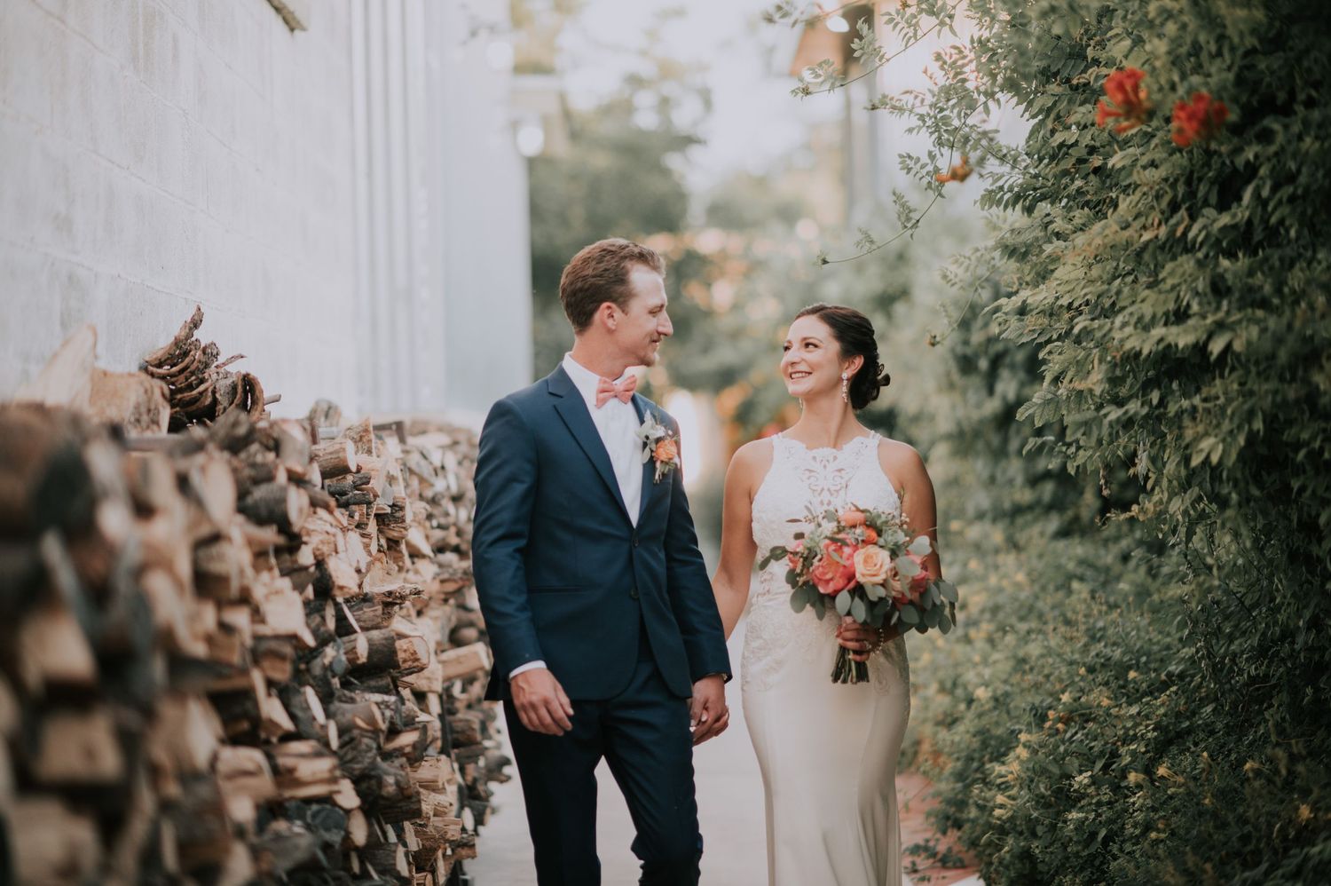 A couple poses together near a stone wall with greenery and flowers.