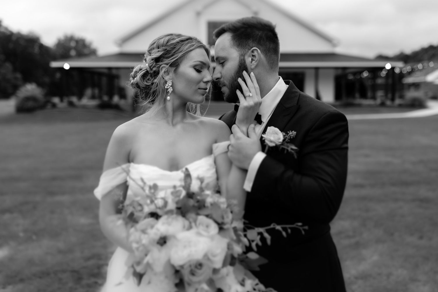 A black and white romantic moment between newlyweds holding a bouquet against a pastoral backdrop.