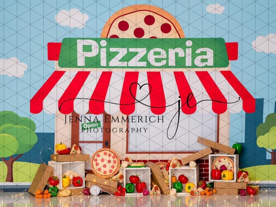 Colorful cartoon pizzeria storefront display with red and white awning, pizza sign, and wooden crates filled with produce.