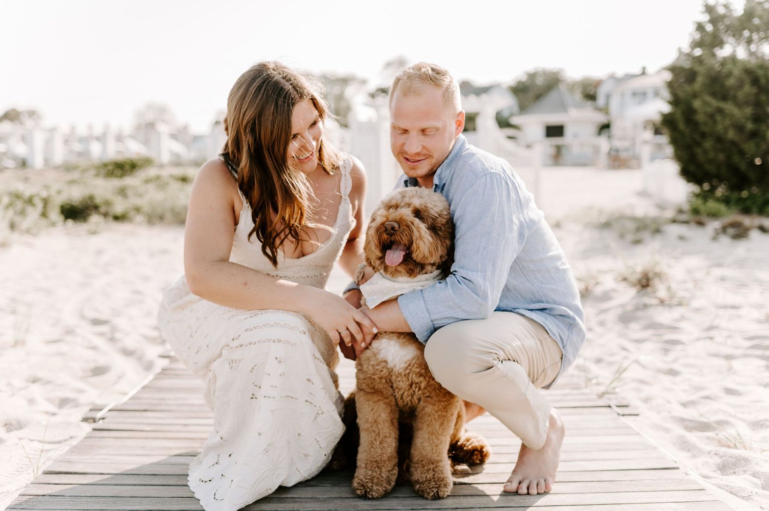 A couple sits on a beach blanket with their goldendoodle dog in casual beachwear.