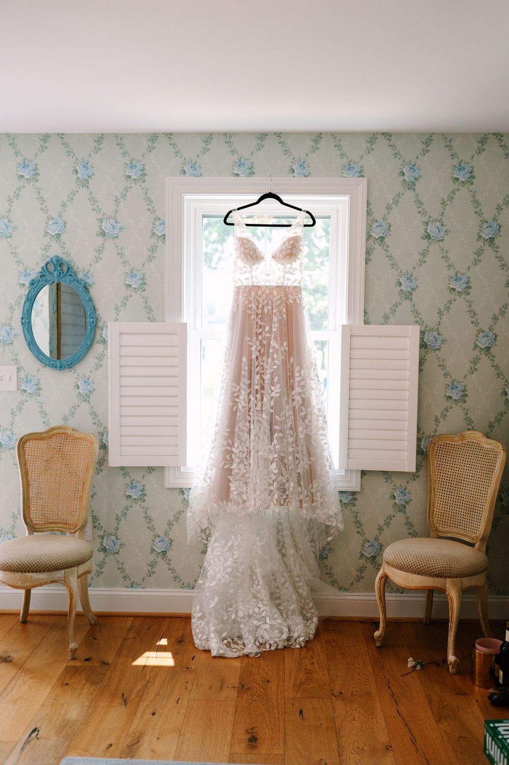 Elegant lace wedding dress hanging in a room with vintage chairs and blue floral wallpaper.