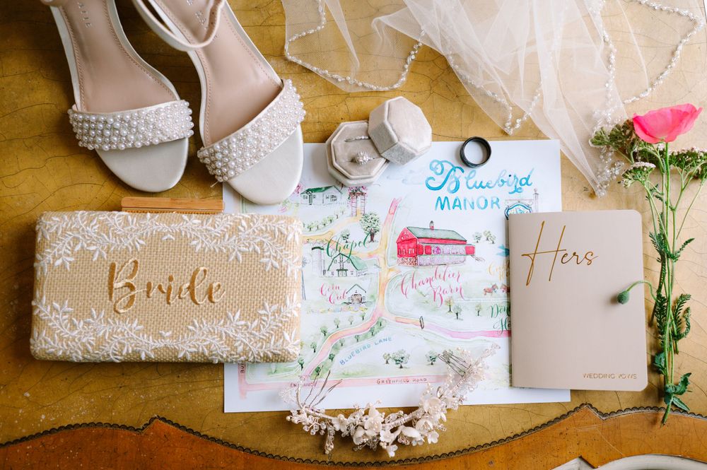 Wedding accessories including pearl shoes, bride clutch, and invitation cards on rustic surface.