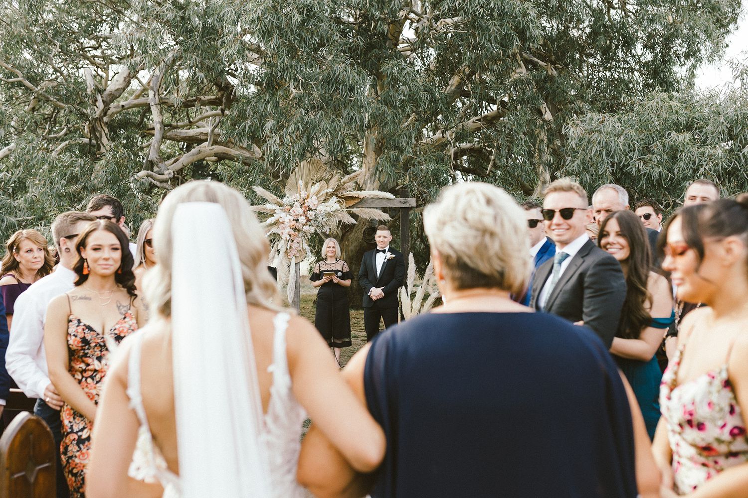 Outdoor wedding ceremony with guests gathered around a natural floral arch under eucalyptus trees.