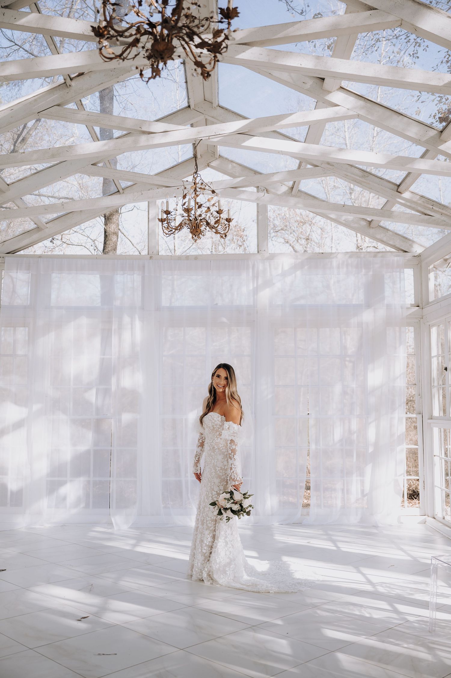 Elegant bridal portrait in white glass conservatory with crystal chandeliers and draped fabric.