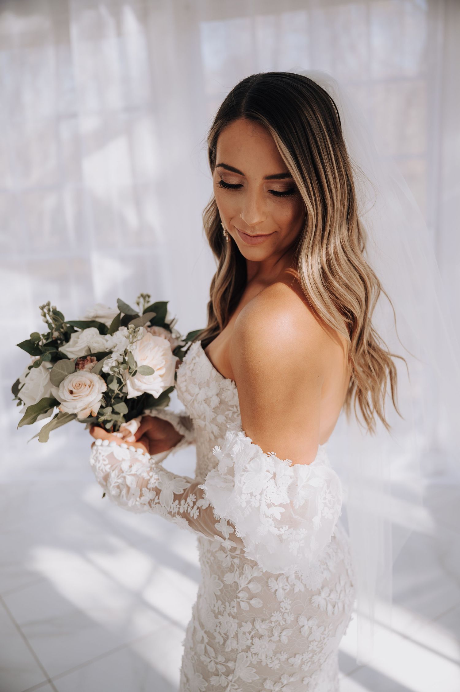 Bride in delicate lace wedding dress holding a white rose bouquet against a sunlit window backdrop.