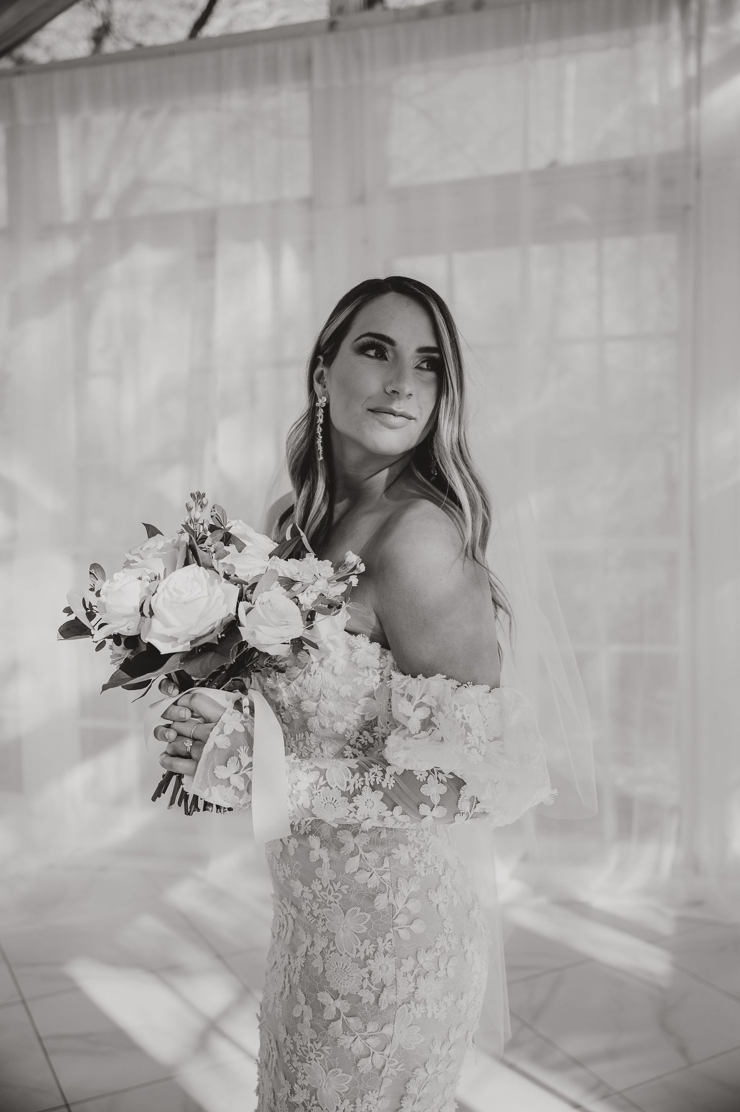 Black and white photo of a bride in a lace off-shoulder gown holding a white floral bouquet.