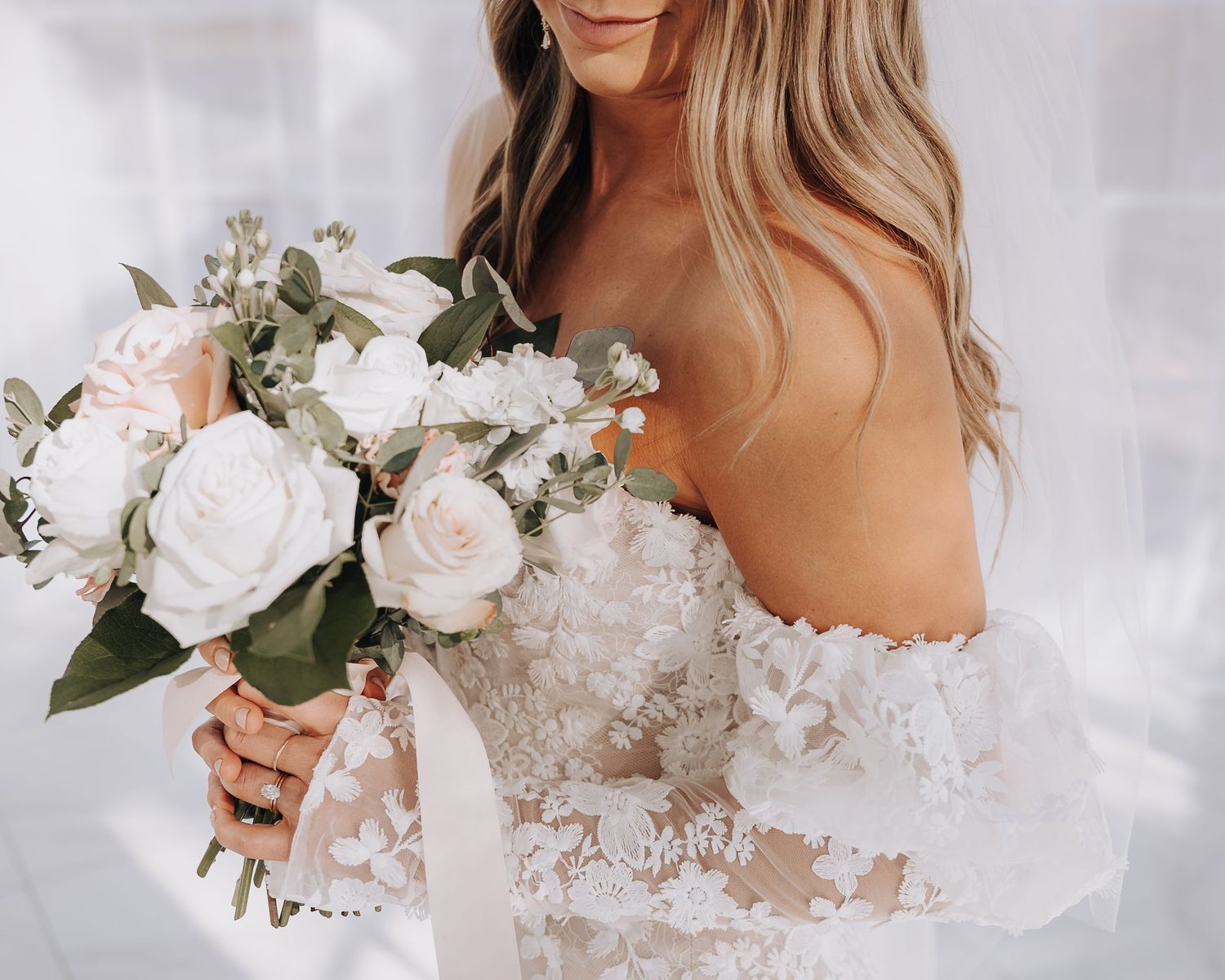 Close up of a bridal bouquet with white roses and lace details on an off-shoulder wedding dress.