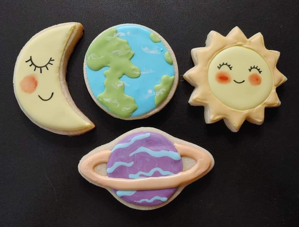 Four decorated sugar cookies of a moon, Earth, sun and planet with a black background.