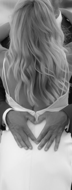 Black and white photo of a groom forming a heart shape with his hands on the back of his bride in a white wedding dress.