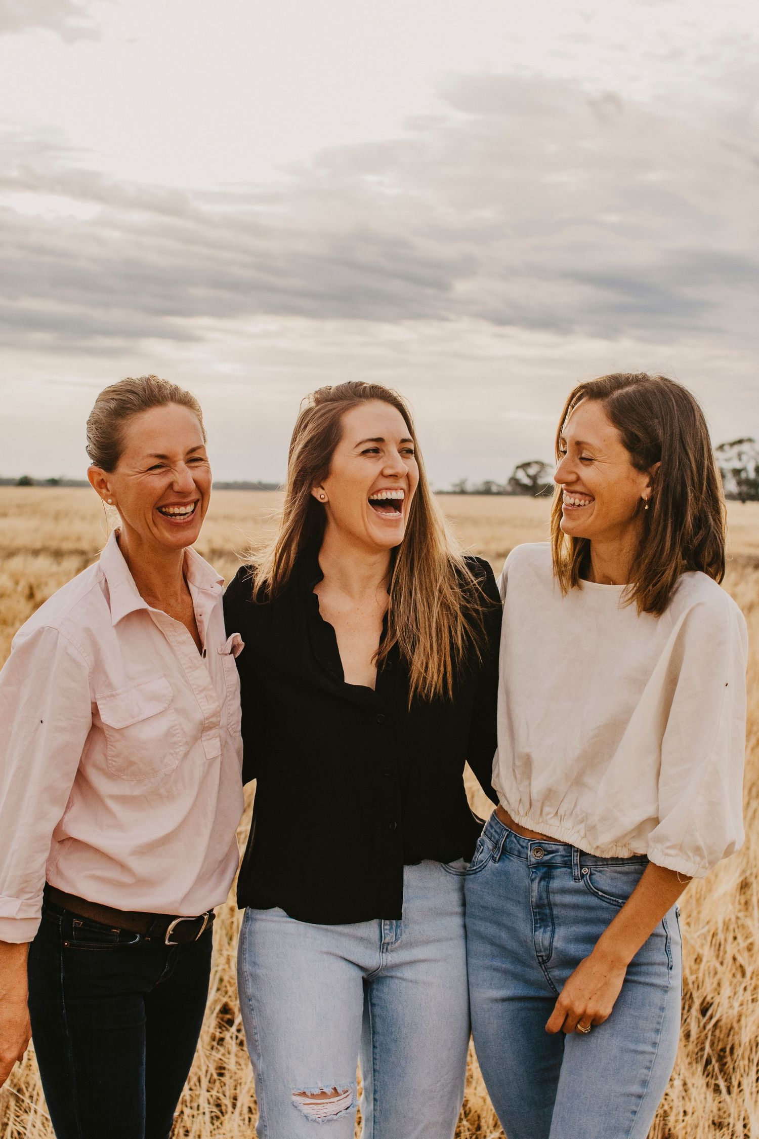 3 rural woman together laughing in a paddock with the sunshine behind them.