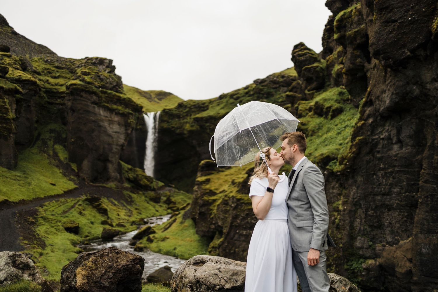Couple with clear umbrella standing by waterfall in mossy volcanic landscape.