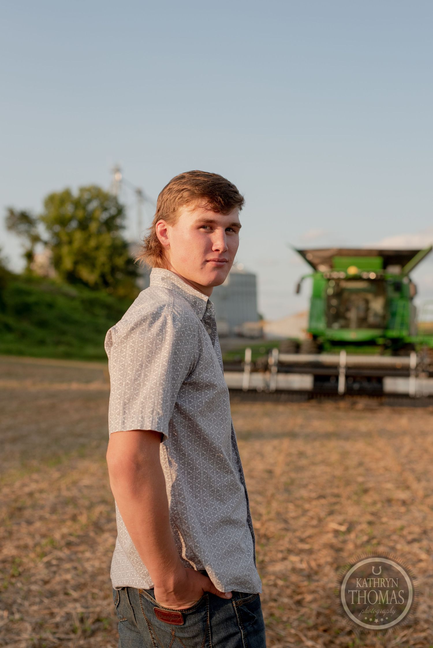 Rural farm scene with agricultural equipment and grain storage silos in background at sunset.