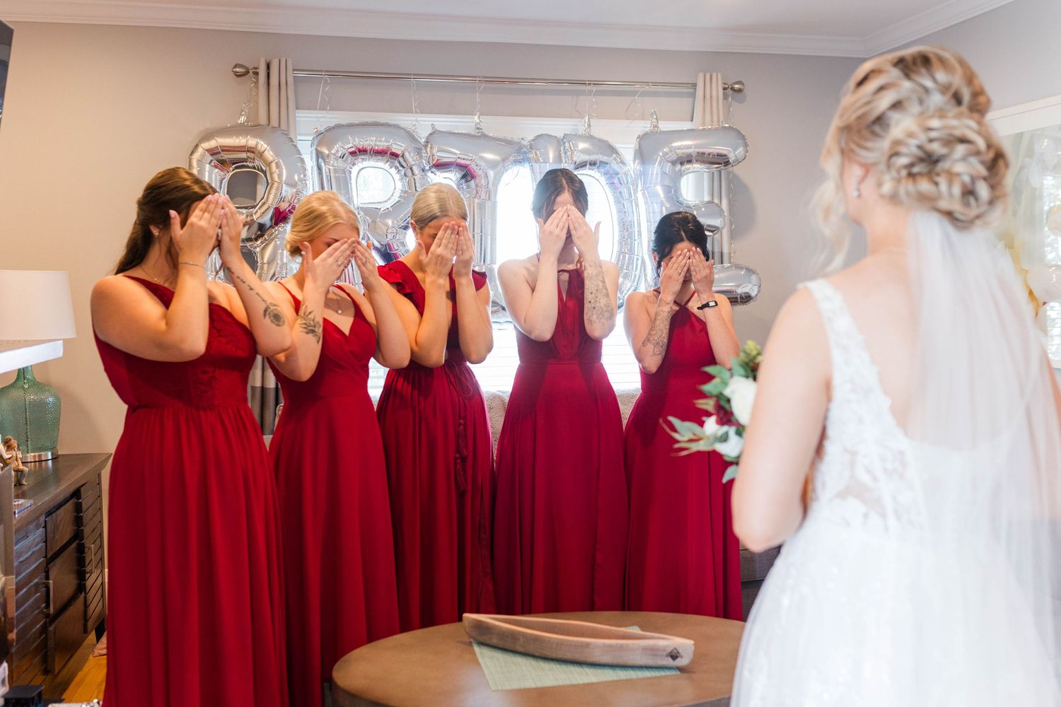 Bridesmaids in flowing red dresses share an emotional first look reaction with bride in white gown.
