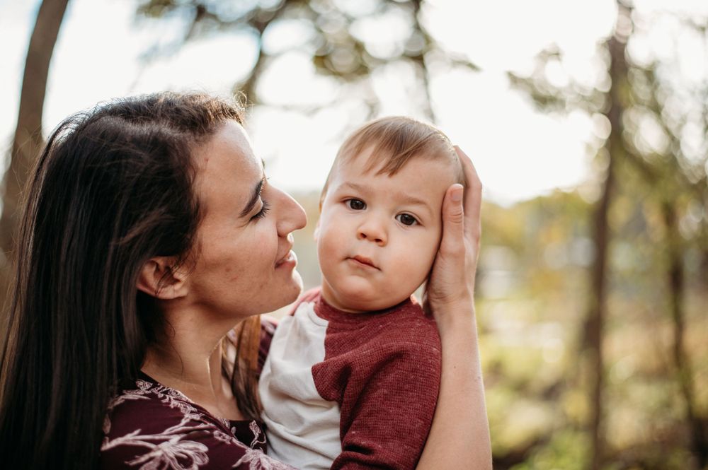 A young toddler smiles directly at the camera while his mom caresses his faces and gives him a kiss on the cheek.