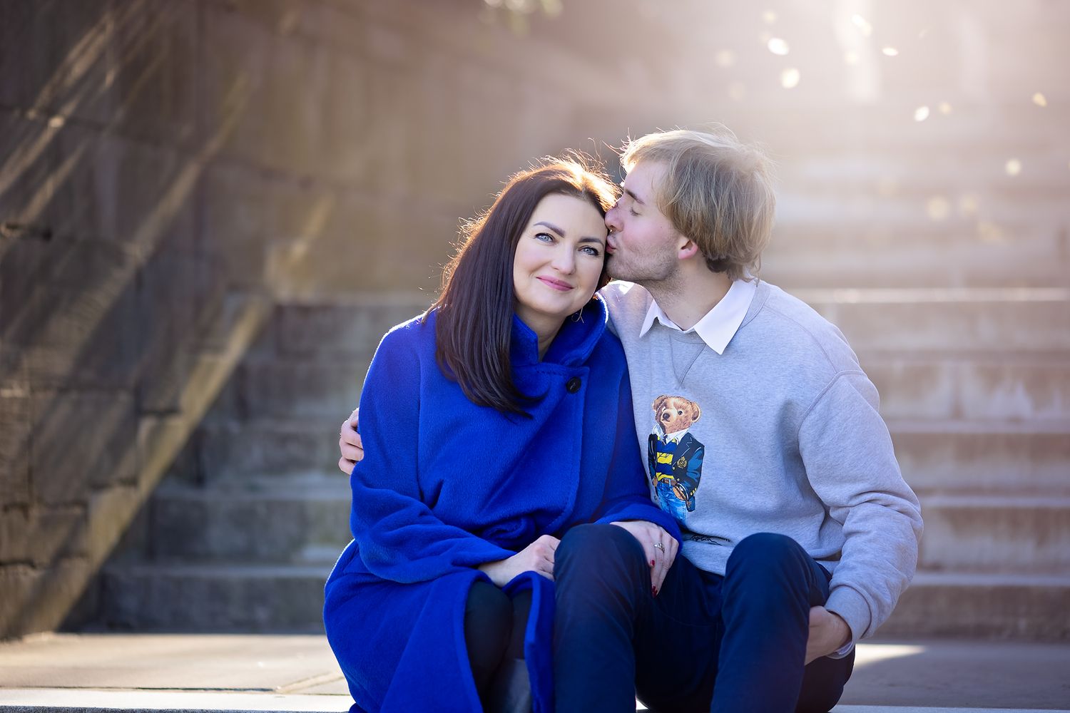 Couple sitting on stone steps outdoors, man kissing woman on cheek in warm sunlight.