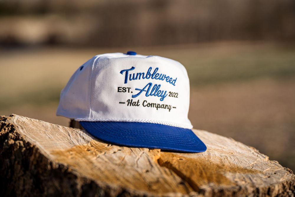 White and blue baseball cap with Tumbleweed Alley logo sitting on wood stump.