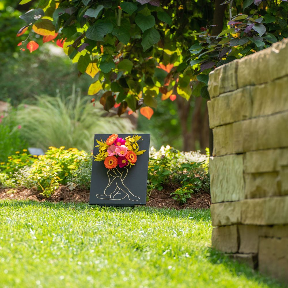 Black canvas floral artwork displayed in lush garden setting with stone wall backdrop.