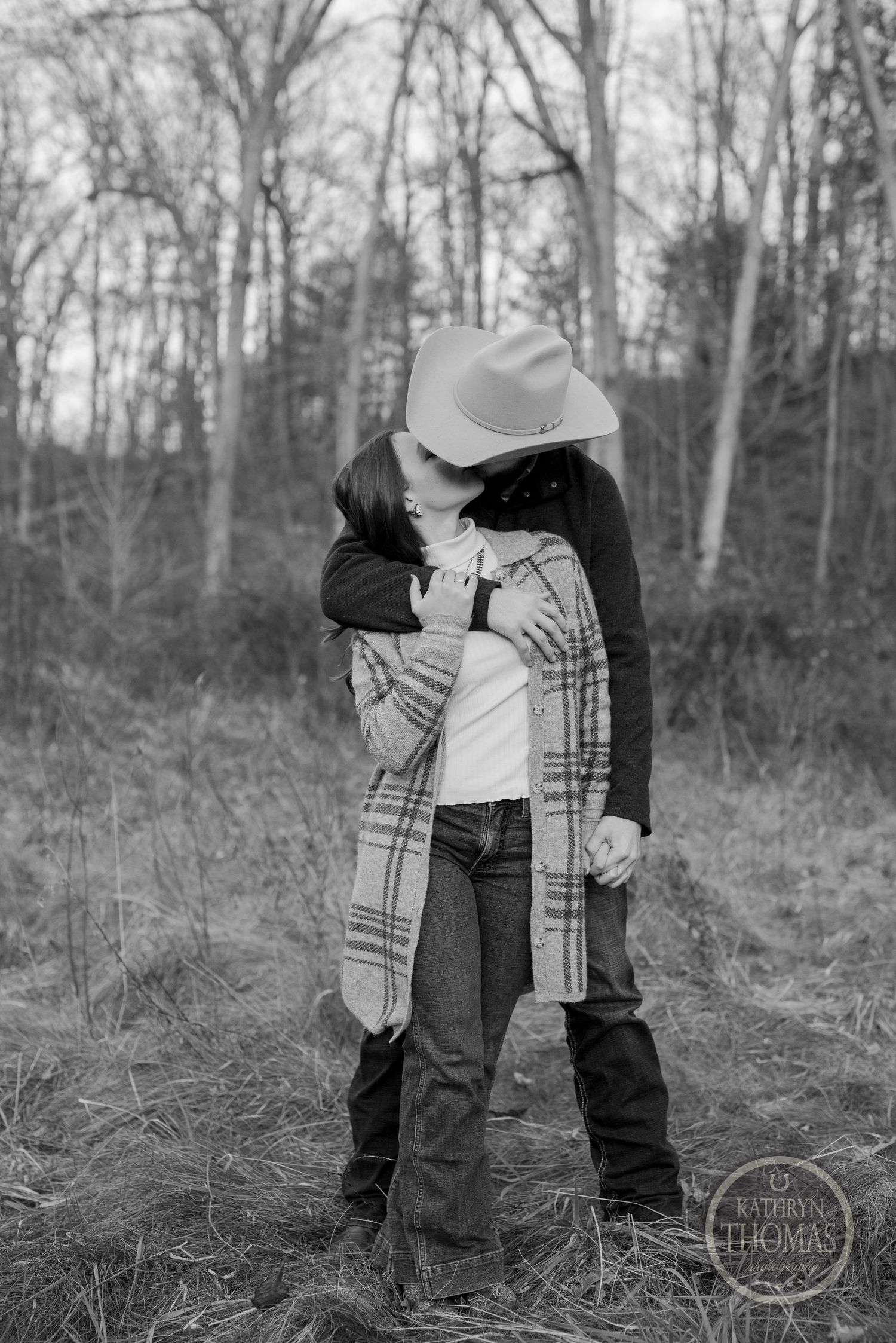 A romantic black and white photo of a couple embracing in a winter forest setting wearing casual clothes and a cowboy hat.