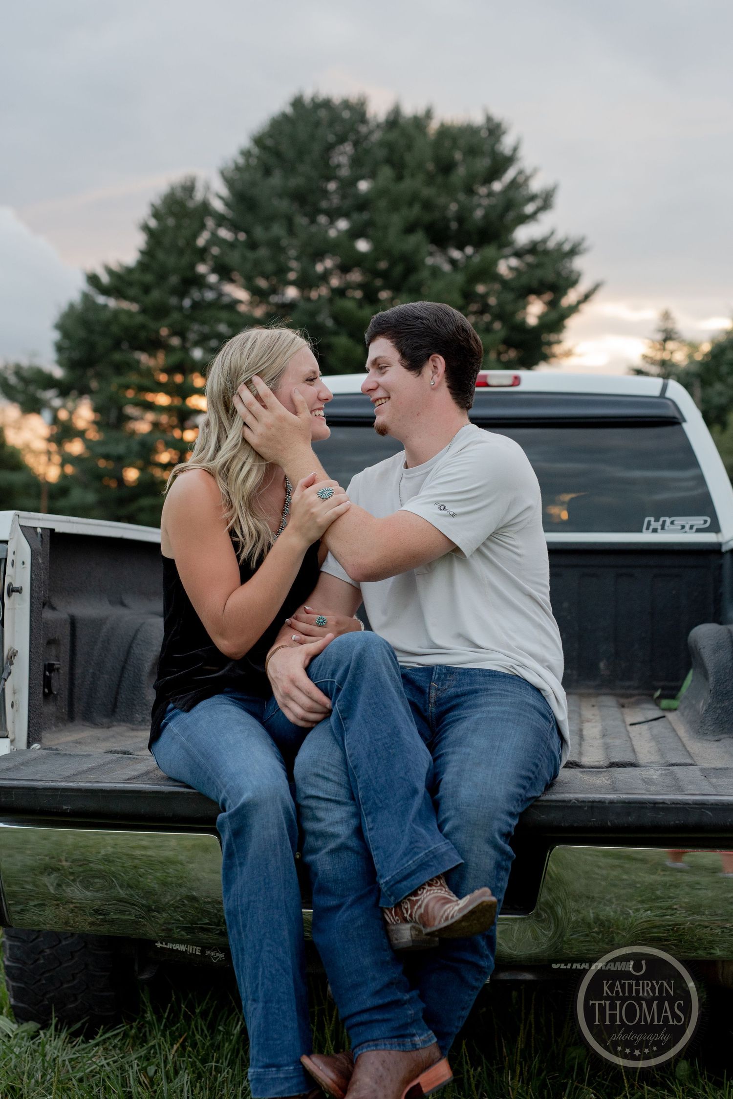 A romantic couple sits in the bed of a pickup truck at sunset with pine trees in the background.