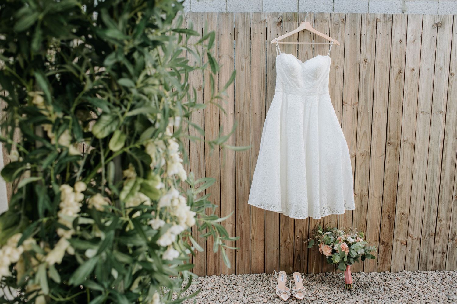 White wedding dress hanging on wooden fence with floral bouquet and greenery.