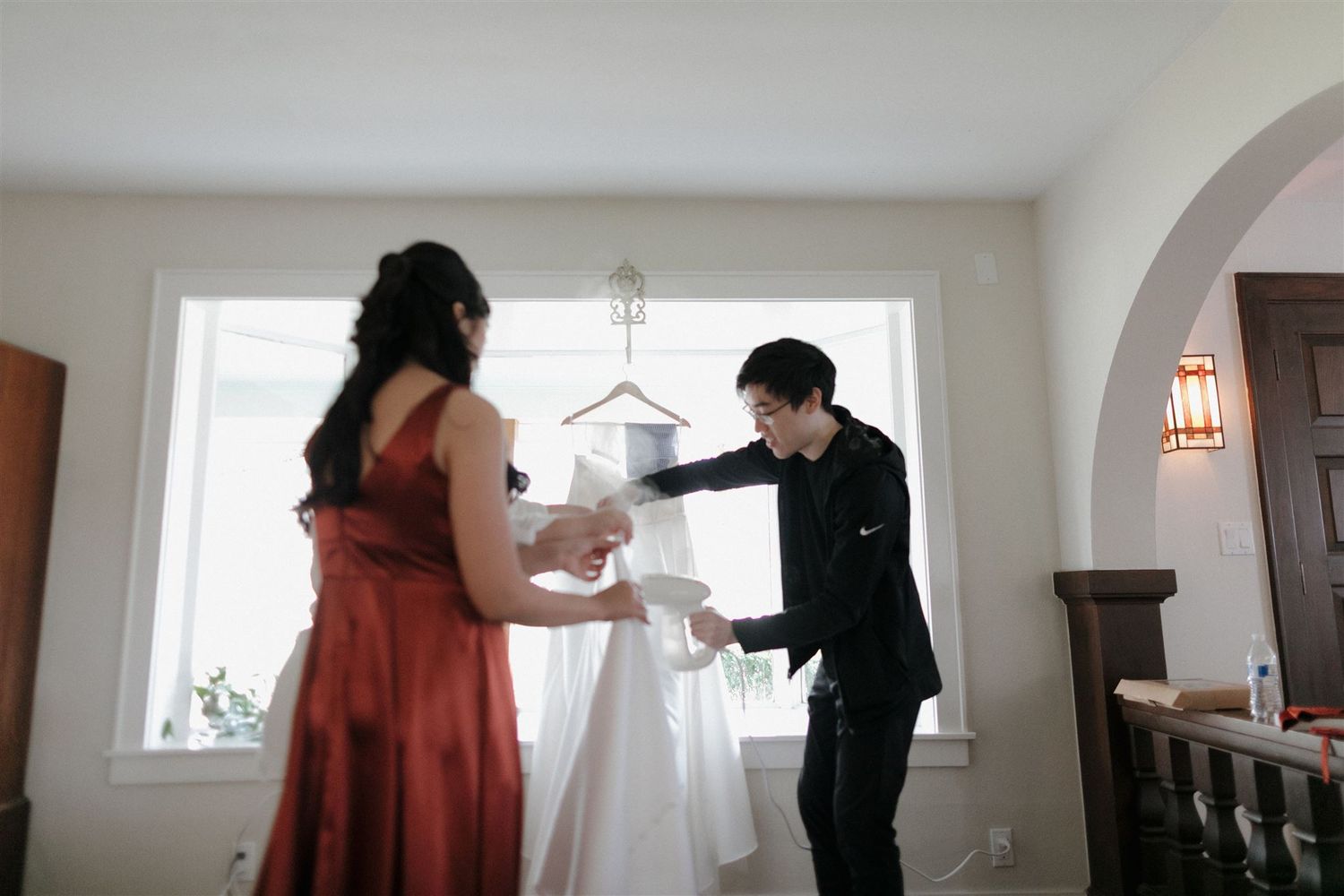 Two people examine and adjust a white wedding gown hanging by a bright window.